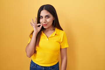 Young arab woman standing over yellow background mouth and lips shut as zip with fingers. secret...