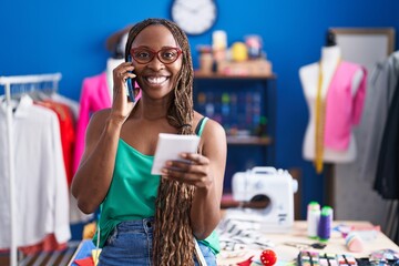 African american woman tailor talking on smartphone reading notebook at atelier