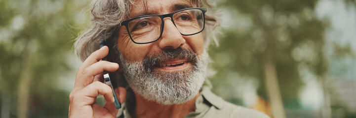 Panorama, Close up portrait od friendly middle aged man with gray hair and beard wearing casual clothes using his mobile phone. Mature gentleman in eyeglasses talking on cell phone outdoors - obrazy, fototapety, plakaty