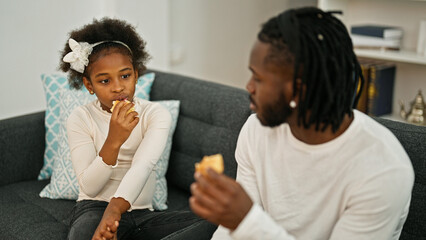 African american father and daughter sitting on sofa eating croissant at home