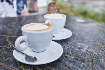 Two cappuccinos on marble table at outdoor cafe, with focus on the creamy froth and inviting ambiance.