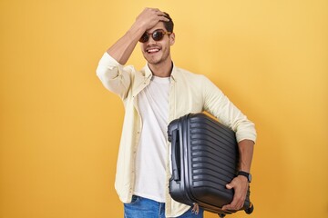 Young hispanic man holding suitcase going on summer vacation surprised with hand on head for...