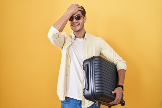 Young hispanic man holding suitcase going on summer vacation surprised with hand on head for mistake, remember error. forgot, bad memory concept.