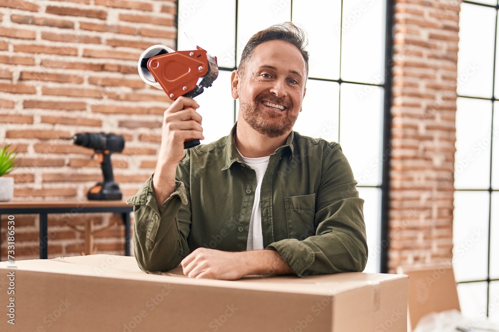 Sticker young caucasian man smiling confident packing cardboard box at new home