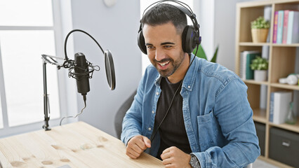 Smiling hispanic man with headphones in a radio studio facing a microphone.