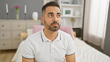 Pensive hispanic man with beard sitting in a modern bedroom with neutral tones