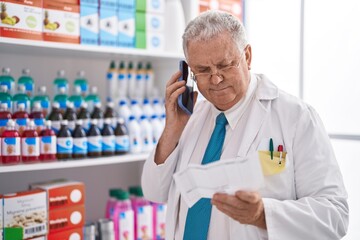 Middle age grey-haired man pharmacist talking on smartphone reading prescription at pharmacy