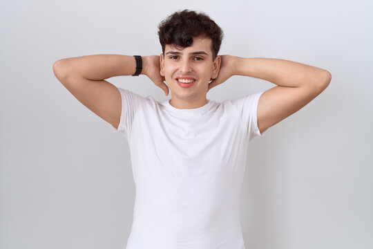 Young non binary man wearing casual white t shirt relaxing and stretching, arms and hands behind head and neck smiling happy