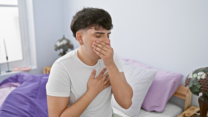 A young man feeling unwell, covering his mouth, sits in a modern bedroom with a simple decor.