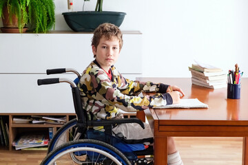 A young boy in a wheelchair concentratedly sits at a desk, engaged in doing his homework.
