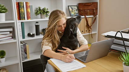 A young woman affectionately embracing her black labrador while working on a laptop in a modern...
