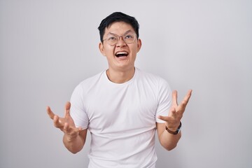 Young asian man standing over white background crazy and mad shouting and yelling with aggressive expression and arms raised. frustration concept.