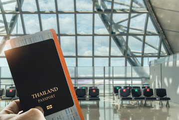 Close-up of a traveler's hand holding a Thai passport in a bright airport terminal, with empty...