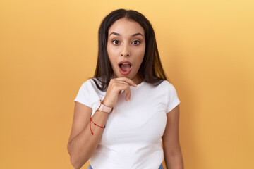 Young arab woman wearing casual white t shirt over yellow background looking fascinated with disbelief, surprise and amazed expression with hands on chin