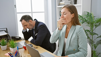 Two exhausted business workers yawning together, signifying long hours of collaborative work at...