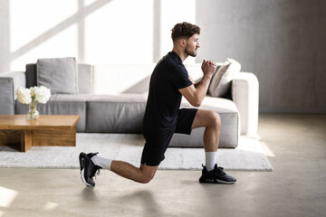 Handsome young man in sportswear with fitness tracker, squats and looks at laptop in living room