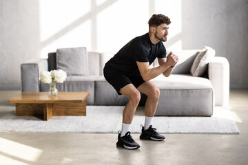 Handsome young man in sportswear with fitness tracker, squats and looks at laptop in living room