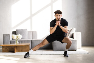 Fototapeta premium Handsome young man in sportswear with fitness tracker, squats and looks at laptop in living room