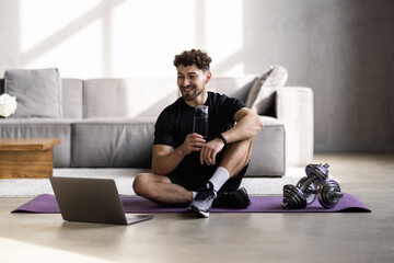 Handsome man using laptop and drinking water while having break during workout at home