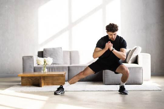 Handsome young man in sportswear with fitness tracker, squats and looks at laptop in living room