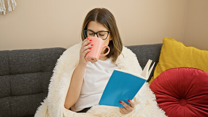 A young hispanic woman enjoys reading and sipping tea, relaxing on a sofa in a cozy living room.