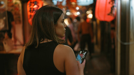 Beautiful hispanic woman's back view as she walks away in tokyo's omoide yokocho, smartphone in...