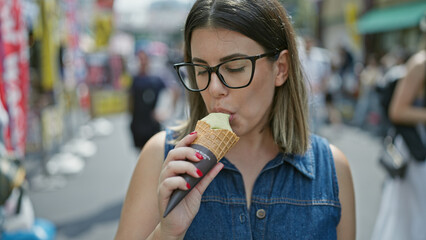 Cheerful and beautiful hispanic woman in glasses happily savors a delicious ice cream cone amidst...