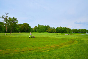 A golf course, with meticulous lawn care underway as a lawnmower trims the green expanses,