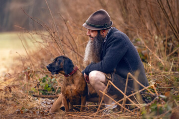 A bavarian man wearing a traditional folk costume interacting with his bavarian mountain dog nearby