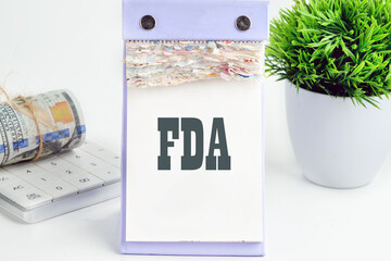 Word FDA written on a desktop tear-off calendar on a white background, next to a calculator with a roll of banknotes with a flower out of focus in the background