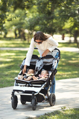 Vertical photo lifestyle young casual caucasian mother take care of two little twin girls in a double baby stroller, walking in city the park	
