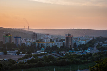 Urban sunset with cityscape, smokestack emissions, and vibrant sky over residential buildings and rolling hills