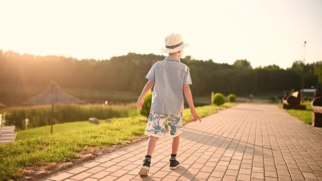 Boy in a hat walks along the sidewalk near a lake and forest on a summer day at sunset. Boy dreams of looking into the distance. Childhood, solitude with nature, dreams.