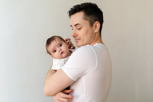 Positive young father in white t shirt admiring and tenderly carrying adorable little baby while looking at her and standing against gray wall background