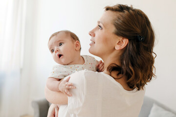 Side view of cheerful young brunette woman in casual clothes embracing precious infant child in...