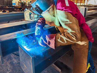 Welder Welding Steel Plate for Steel Structure Work with Process Flux Cored Arc Welding (FCAW) and Appropriately Dressed with Personal Protective Equipment (PPE) for Safety, at an Industrial Factory.