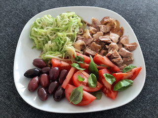 Homemade grilled chicken with zucchini, ovives, sliced tomato and basil leaves, served on a white plate