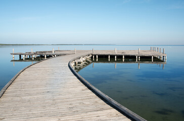 A wide jetty made of wooden planks on shallow water on the shore. The horizon with blue sky in the background.