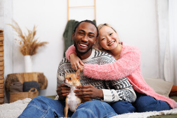 Smiling young diverse couple looking at camera and hugging near pet on bed at home