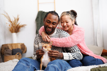 Smiling asian woman hugging african american boyfriend with pet on bed at home