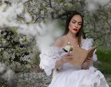 Spring sexy fantasy woman sits on branch of green tree blooming sakura flowers garden. Medieval girl vintage lady. pink silk fly in wind dress. fairy tale image art photo. Brunette long hair cute face