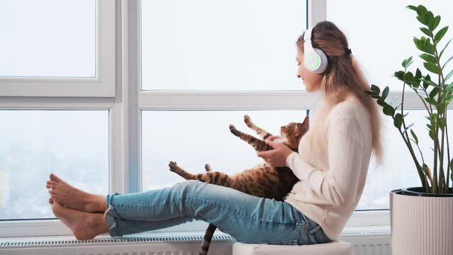 happy blonde woman sits on windowsill panoramic window headphones, listens cheerful driving music with cat animal dances waves paws. Joyful girl fashion model enjoys sound dance with funny disco pet