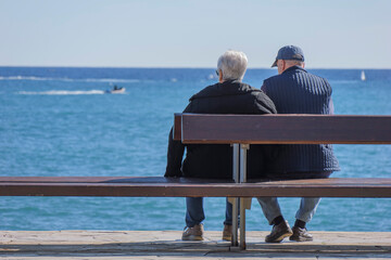Elderly couple sitting on a wooden bench facing the blue Mediterranean Sea, back to back