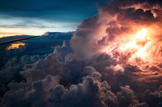 Vista da asa de um avião comercial passando diretamente entre a tempestade e a nuvem Cumulonimbus com raios e trovões. Tons fortes de roxo, azul e laranja.