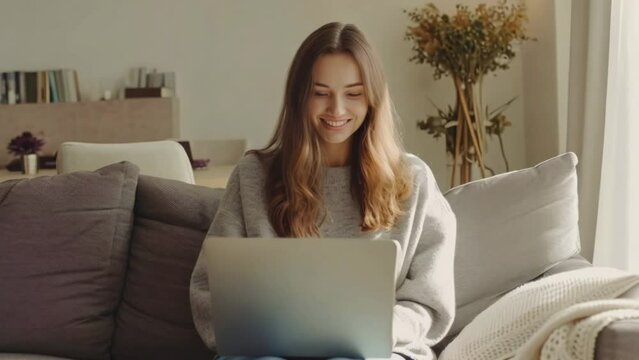 Young freelancer woman sit white sofa home. Person looking at laptop screen. Girl watch video on computer, playing game, or learning online. Distance Internet education concept. Virtual freelance work
