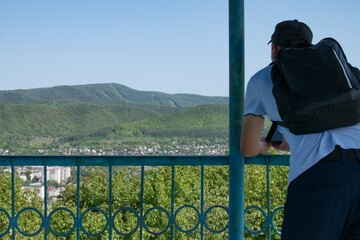 A man in a cap with a backpack stands on the observation deck and looks at the city and the mountains