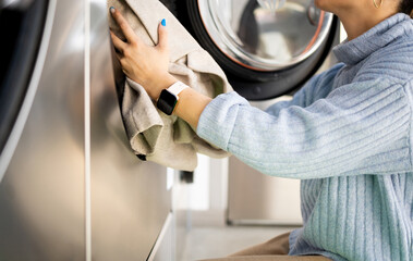 An unrecognizable woman is putting a garment inside a public self-service washing machine. Public...