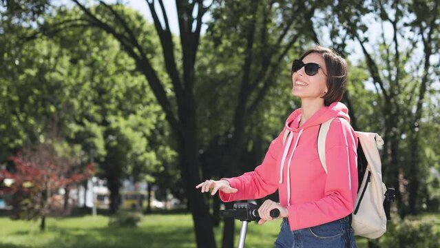 Side view of smiling female with white backpack and sunglasses dressed in bright clothes showing hello gesture outdoors. Pretty caucasian woman having rest after long trip on modern electic scooter.