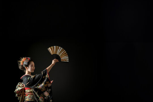 Magnifique Geisha v&ecirc;tue d'un kimono en soie traditionnel et d'une coiffure ancestrale avec des peignes &agrave; chignon ex&eacute;cutant la danse de l'&eacute;ventail sur fond noir avec espace n&eacute;gatif pour texte copyspace