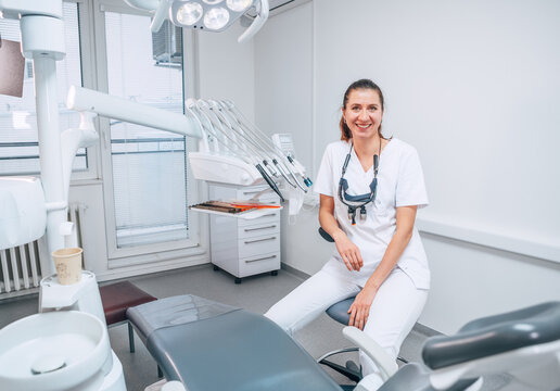 Portraait of sincerely smiling young dentist woman dressed white medical scrubs uniform sitting in modern dental clinic next to stomatology  chair. Health care and medicare industry concept image.